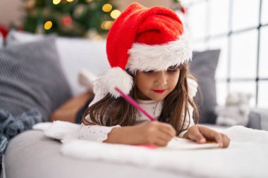 Adorable hispanic girl drawing on notebook lying on sofa by christmas tree at home