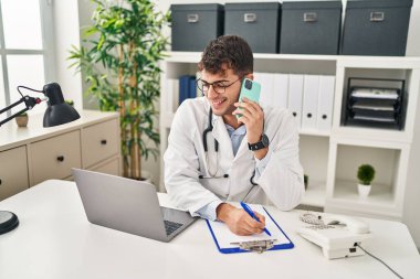 Young hispanic man doctor talking on smartphone writing medical report at clinic
