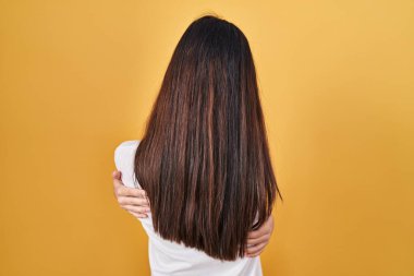 Young beautiful woman standing over yellow background hugging oneself happy and positive from backwards. self love and self care 