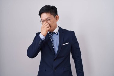 Young asian man wearing business suit and tie smelling something stinky and disgusting, intolerable smell, holding breath with fingers on nose. bad smell 