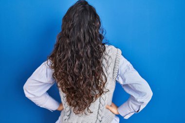 Genç esmer kadın mavi arka planda dikiliyor geriye bakıyor kolları vücudunda. 