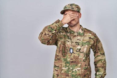 Young hispanic man wearing camouflage army uniform smiling and laughing with hand on face covering eyes for surprise. blind concept. 