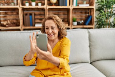 Orta yaşlı beyaz bir kadın evdeki kanepede otururken akıllı telefon kullanıyor..