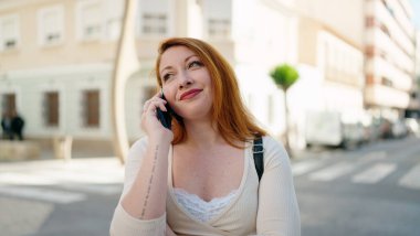 Genç kızıl saçlı, gülümseyen, kendine güvenen, caddedeki akıllı telefondan konuşan kadın.