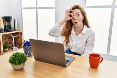 Young caucasian woman working at the office using computer laptop doing ok gesture shocked with surprised face, eye looking through fingers. unbelieving expression. 