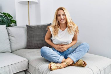 Beautiful blonde woman sitting on the sofa at home with hand on stomach because nausea, painful disease feeling unwell. ache concept. 
