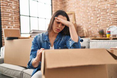 Young beautiful hispanic woman unpacking cardboard box with worried expression at new home