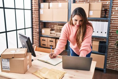 Young beautiful hispanic woman ecommerce business worker using laptop writing on notebook at office