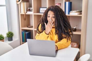 Plus size hispanic woman using laptop at home covering mouth with hand, shocked and afraid for mistake. surprised expression 