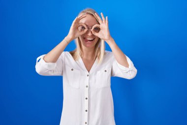 Mavi arka planda dikilen genç beyaz kadın dürbün gibi jest yapıyor. Dürbün gibi dilini çıkartıyor, gözler parmaklarından bakıyor. çılgın ifade. 