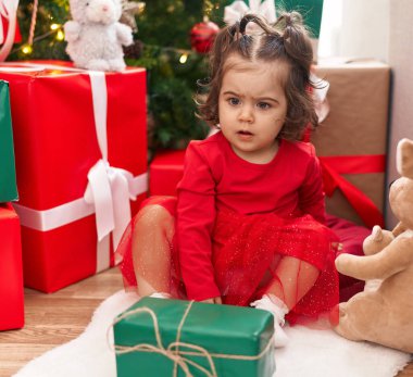 Adorable hispanic girl sitting on floor by christmas tree at home