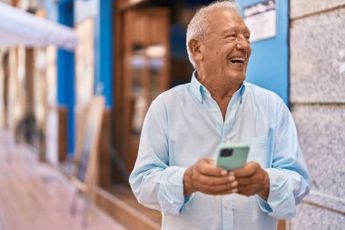 Yaşlı, gri saçlı, kendine güvenen, caddede akıllı telefon kullanan bir adam.