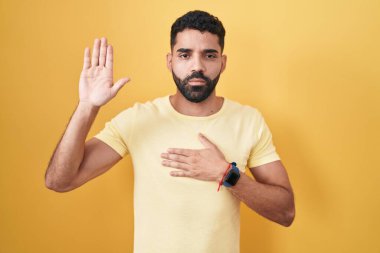 Hispanic man with beard standing over yellow background swearing with hand on chest and open palm, making a loyalty promise oath 