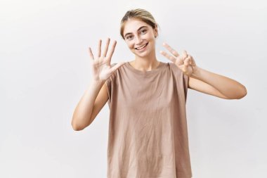 İzole edilmiş arka planda duran genç sarışın kadın yedinci parmaklarını gösterip işaret ederken kendinden emin ve mutlu bir şekilde gülümsüyor.. 