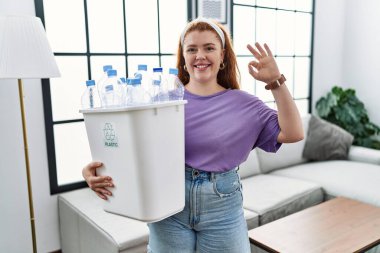 Young redhead woman holding recycling wastebasket with plastic bottles smiling positive doing ok sign with hand and fingers. successful expression. 