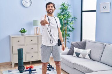 Handsome latin man wearing sportswear at home smiling friendly offering handshake as greeting and welcoming. successful business. 