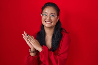 Asian young woman standing over red background clapping and applauding happy and joyful, smiling proud hands together 