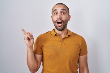 Hispanic man with beard standing over white background surprised pointing with finger to the side, open mouth amazed expression. 