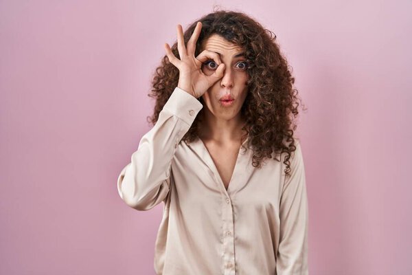 Латиноамериканка с вьющимися волосами, стоящими на розовом фоне и делающими нормальный жест, шокированная удивленным лицом, глазами, смотрящими сквозь пальцы. неверное выражение. 