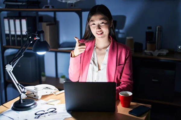 Chinese young woman working at the office at night doing happy thumbs up gesture with hand. approving expression looking at the camera showing success.