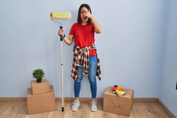 Young hispanic woman painting home walls with paint roller peeking in shock covering face and eyes with hand, looking through fingers with embarrassed expression.