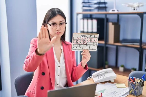 Chinese young woman working at the office holding holidays calendar with open hand doing stop sign with serious and confident expression, defense gesture