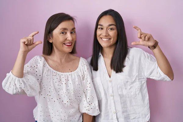 Hispanic mother and daughter together smiling and confident gesturing with hand doing small size sign with fingers looking and the camera. measure concept.