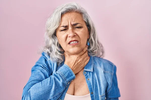 Middle age woman with grey hair standing over pink background touching painful neck, sore throat for flu, clod and infection
