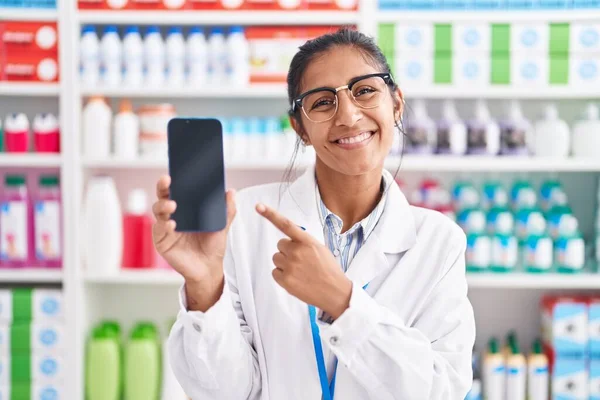 Young hispanic woman working at pharmacy drugstore showing smartphone screen smiling happy pointing with hand and finger