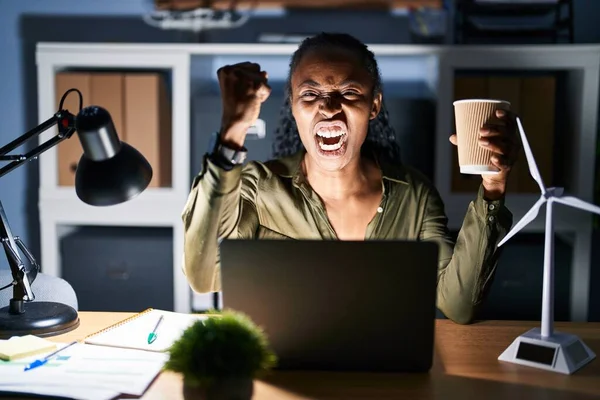African woman working using computer laptop at night angry and mad raising fist frustrated and furious while shouting with anger. rage and aggressive concept.