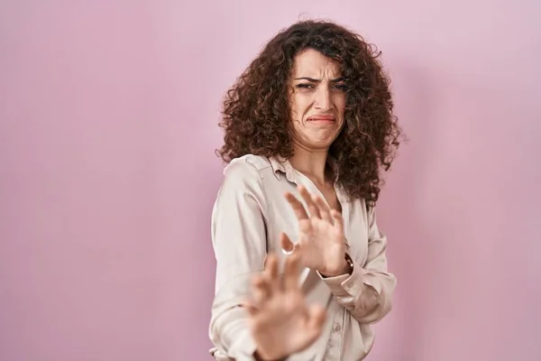 Hispanic woman with curly hair standing over pink background disgusted expression, displeased and fearful doing disgust face because aversion reaction.