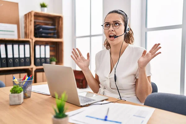 Young hispanic woman wearing call center agent headset at the office crazy and mad shouting and yelling with aggressive expression and arms raised. frustration concept.