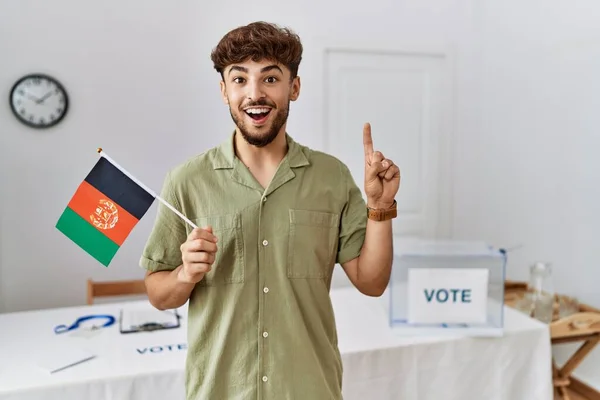 Young arab man at political campaign election holding afghanistan flag surprised with an idea or question pointing finger with happy face, number one