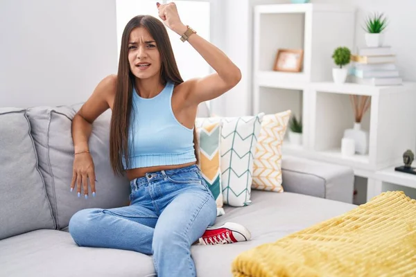 Young brunette woman sitting on the sofa at home angry and mad raising fist frustrated and furious while shouting with anger. rage and aggressive concept.