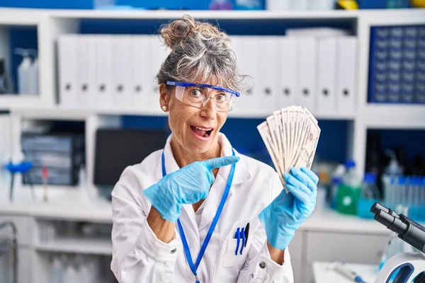 Middle age woman working at scientist laboratory holding dollars smiling happy pointing with hand and finger