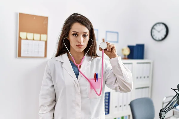 Young doctor woman wearing doctor coat holding stethoscope thinking attitude and sober expression looking self confident