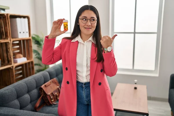Young Latin Woman Working Therapist Holding Jar Pills Pointing Thumb — Stockfoto