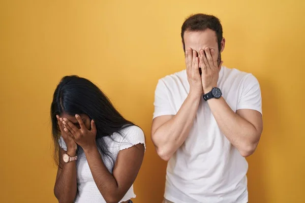 Interracial couple standing over yellow background with sad expression covering face with hands while crying. depression concept.
