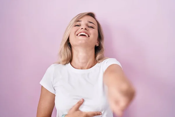 Young blonde woman standing over pink background laughing at you, pointing finger to the camera with hand over body, shame expression