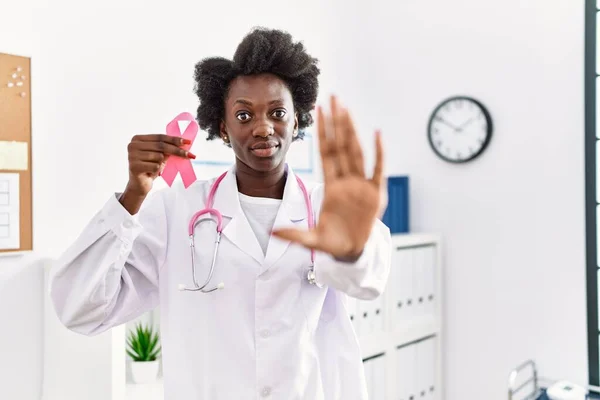 African doctor woman holding pink cancer ribbon at clinic with open hand doing stop sign with serious and confident expression, defense gesture