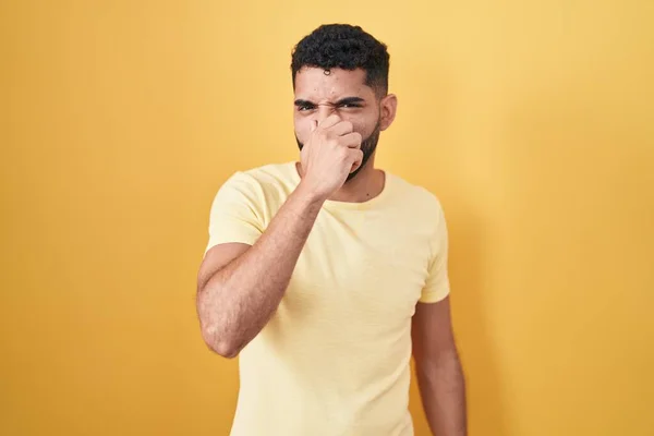 黄色の背景の上に立ってひげを持つヒスパニック系の男は 鼻の上に指で息を保持し 臭いと嫌な 耐え難い匂いを嗅ぐ 臭いが悪い — ストック写真