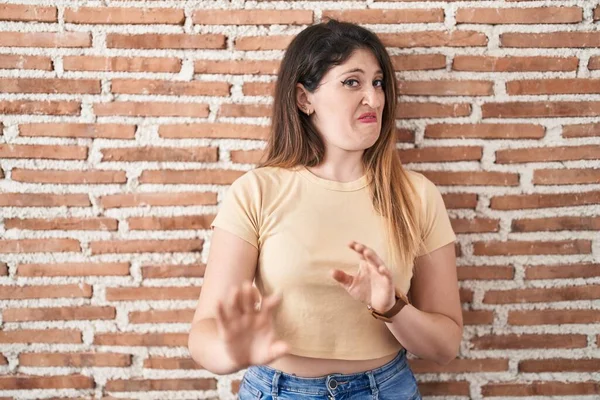 Young brunette woman standing over bricks wall disgusted expression, displeased and fearful doing disgust face because aversion reaction.