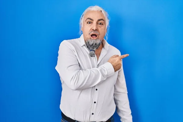 Middle age man with grey hair standing over blue background surprised pointing with finger to the side, open mouth amazed expression.