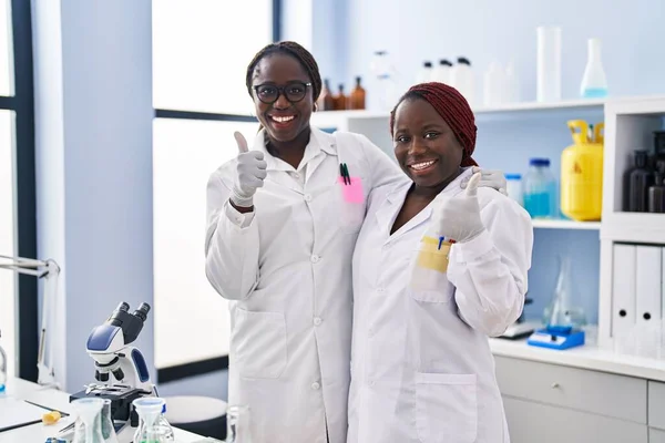 科学研究所で働く2人のアフリカ人女性が笑顔で前向きで 親指を上げて素晴らしい承認サインをします — ストック写真