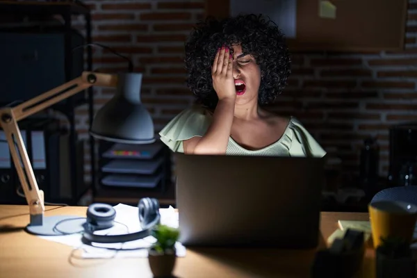 Young brunette woman with curly hair working at the office at night yawning tired covering half face, eye and mouth with hand. face hurts in pain.