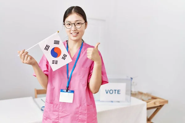 Young asian nurse woman at political campaign election holding south korea flag smiling happy and positive, thumb up doing excellent and approval sign