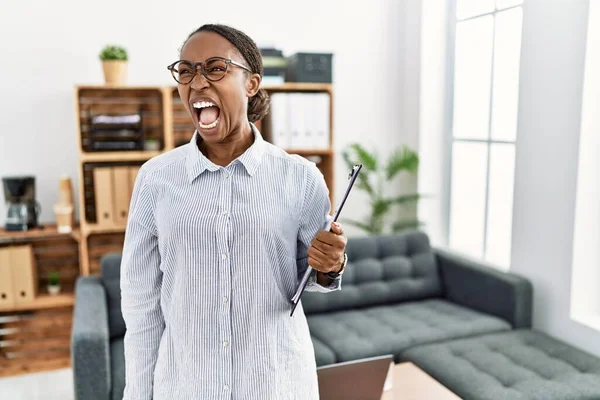 African woman working at psychology clinic angry and mad screaming frustrated and furious, shouting with anger. rage and aggressive concept.