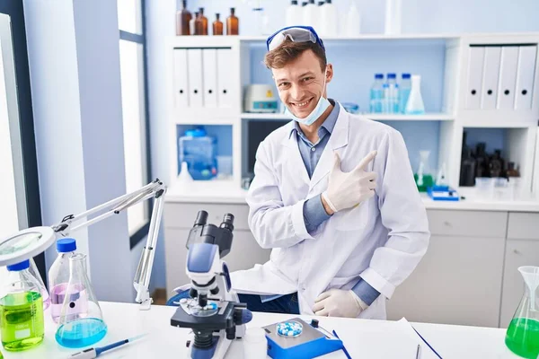 Caucasian man working at scientist laboratory smiling cheerful pointing with hand and finger up to the side