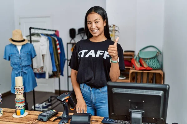 Young hispanic woman working as staff at retail boutique doing happy thumbs up gesture with hand. approving expression looking at the camera showing success.