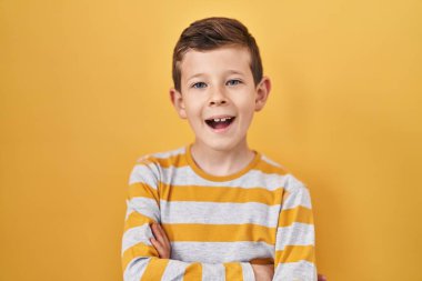 Young caucasian kid standing over yellow background happy face smiling with crossed arms looking at the camera. positive person. 